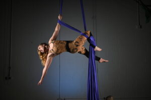 A woman with blond hair and a matching colored unitard performs a trick on purple aerial silks