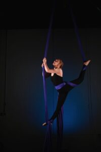 A woman in a black unitard performs a split maneuver on a purple aerial silk