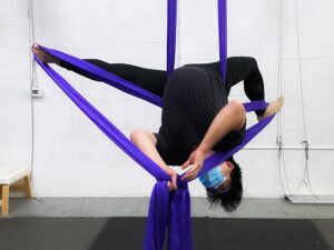 A man in a gray t-shirt and black leggings performs an inverted split pose in a purple aerial silk