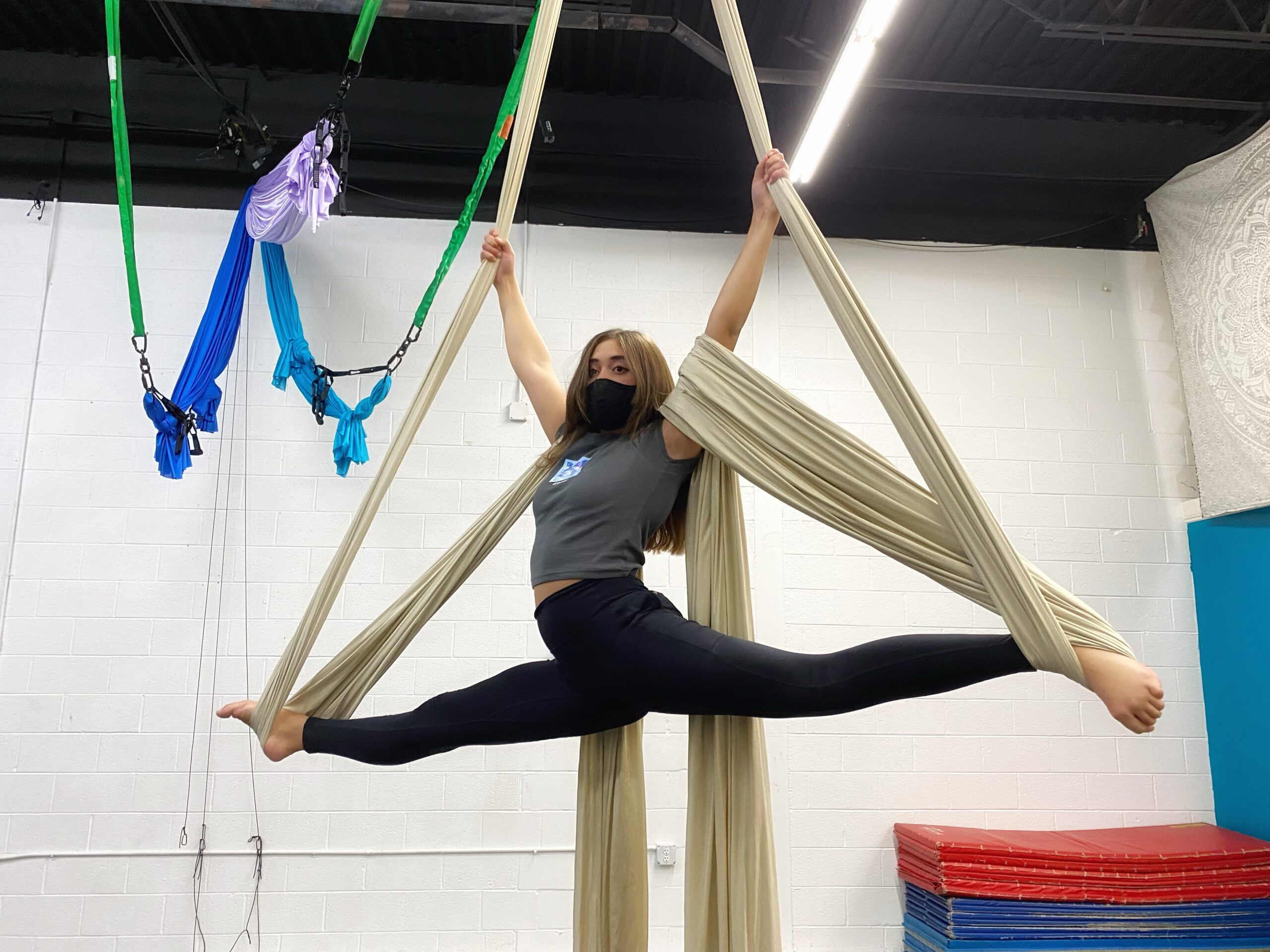 A silks student wearing a gray crop top and black leggings performs a split on champagne colored aerial silks