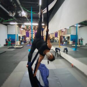 A woman in black tank top and leggings performs a standing middle split on navy blue aerial silks