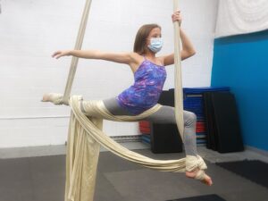 A teen girl in a purple camisole and gray leggings performs a stag split on a champagne colored silk