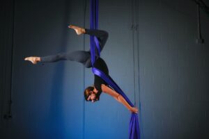 A woman in a black shirt and gray leggings performs a trick on purple aerial silks