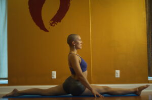 A bald woman wearing a purple sports bra and gray shorts performs a split on a blue yoga mat in front of a gold colored wall